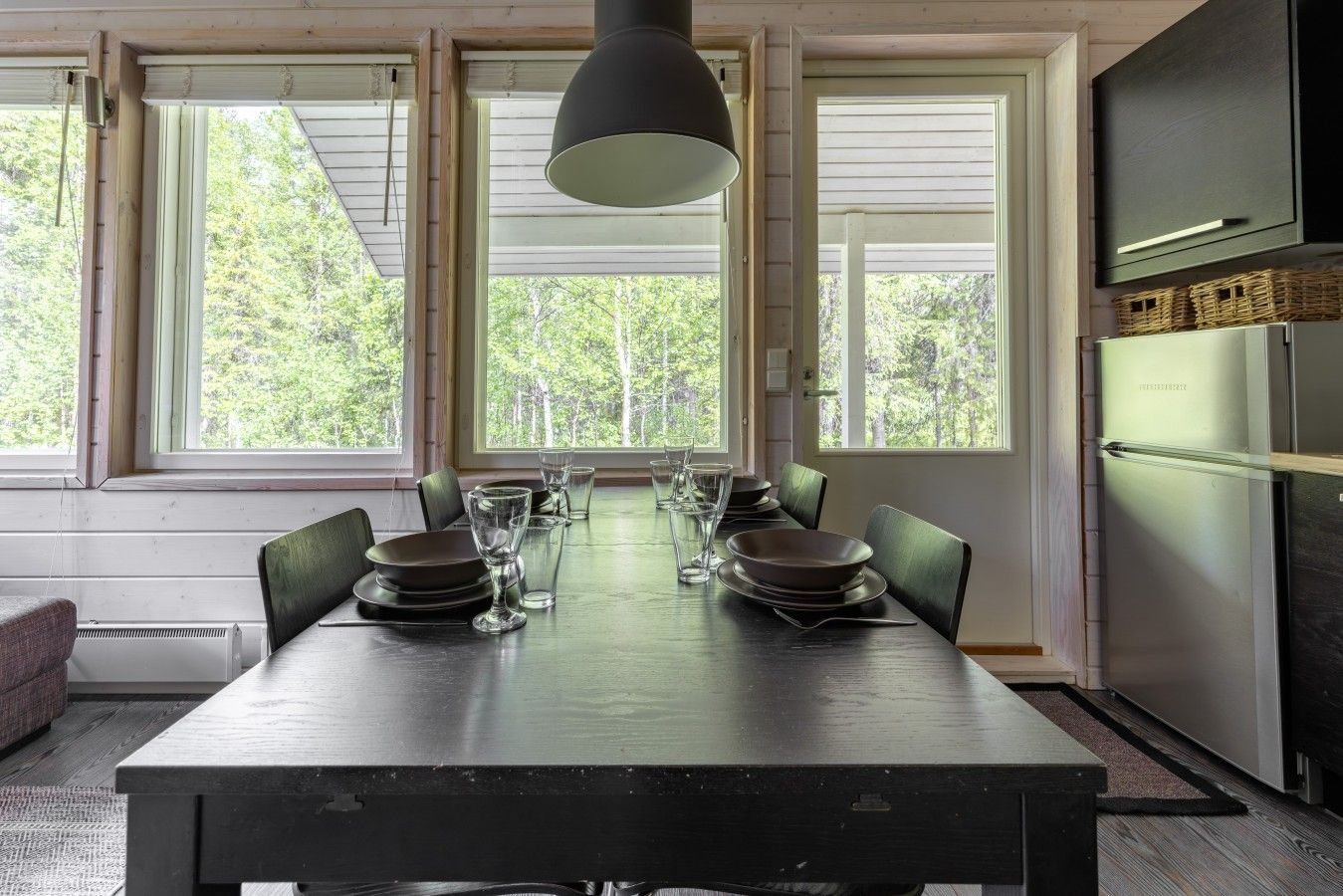 Un coin repas avec une table dressée pour quatre personnes, de grandes fenêtres et une cuisine moderne équipée d’un réfrigérateur.
