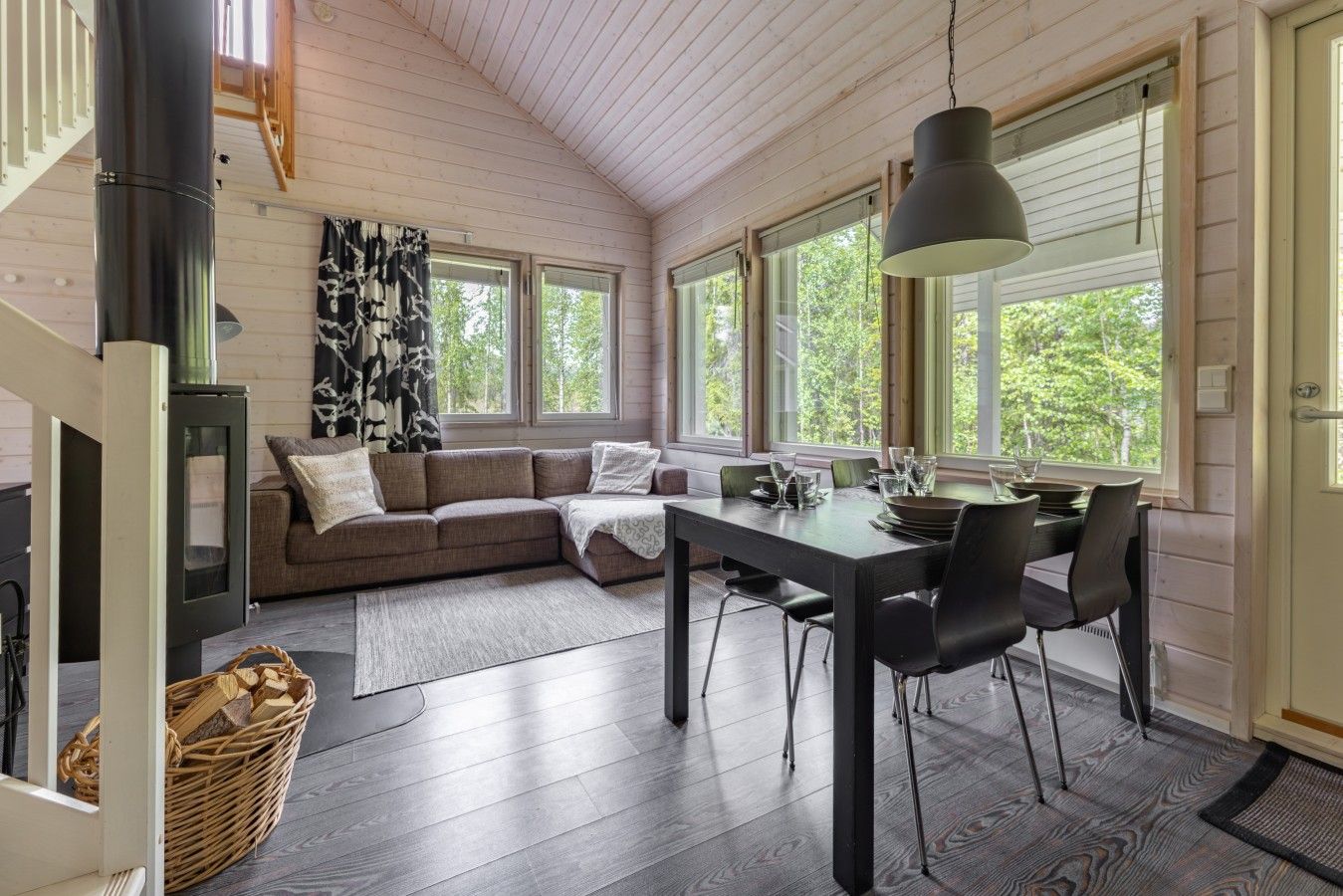 A cozy interior with a wooden ceiling, a sofa, a dining table with chairs, and large windows.