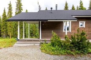 Mökki, jossa on katettu kuisti, puinen ulkoverhous ja sorapäällysteinen ajotie.