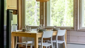 Un espace repas avec une table en bois et des chaises blanches, de grandes fenêtres donnant sur la verdure, et un coin cuisine équipé d’un réfrigérateur et de placards.
