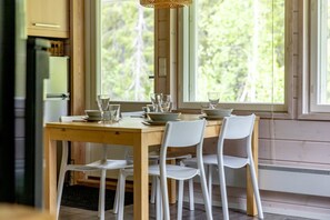 A dining area with a wooden table and white chairs, large windows with a view of trees, and a kitchen area with a refrigerator and cabinets.