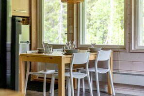 A dining area with a wooden table and white chairs, large windows with a view of trees, and a kitchen area with a refrigerator and cabinets. - Villa Lumi 2 | Cozy Arctic Retreat in Ylläs (Ylläsjärvi)
