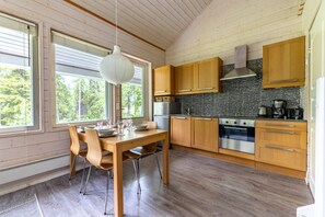 A modern kitchen with wooden cabinets, a stainless steel oven, and a large window with a view of trees. - Villa Lumi 1 | Cozy Arctic Retreat in Ylläs (Ylläsjärvi)