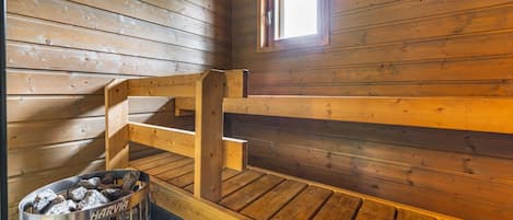 Interior de una cabaña de madera con literas, una ventana y un contenedor metálico con la etiqueta 'HARVIA' en el suelo.