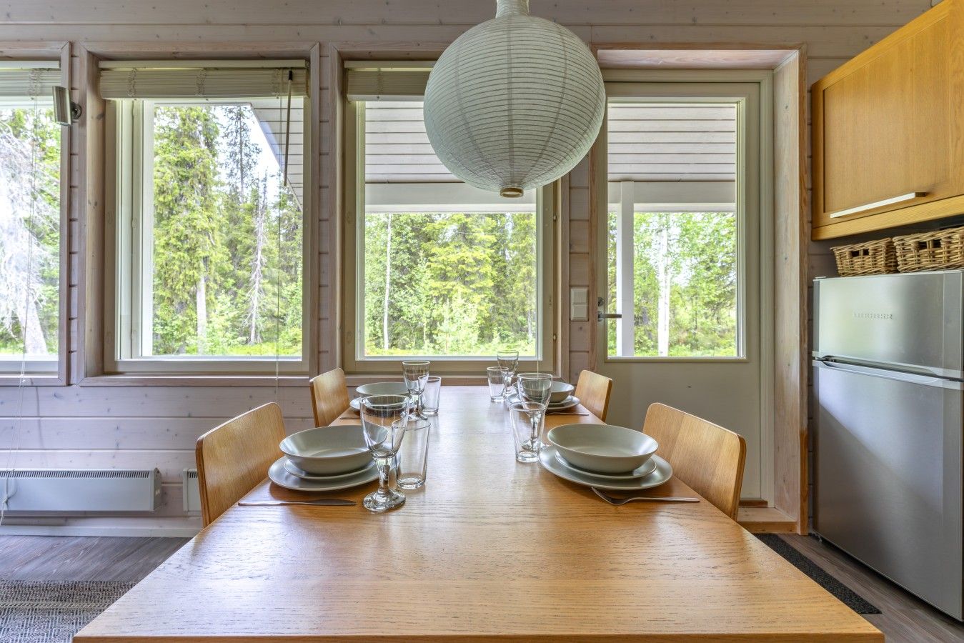 A dining area with a wooden table set for six, large windows, and a modern kitchen with a stainless steel refrigerator.