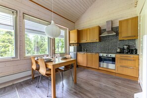 A modern kitchen with wooden cabinets, a stainless steel oven, and a large window with a view of trees. - Villa Lumi 1 | Cozy Arctic Retreat in Ylläs (Ylläsjärvi)
