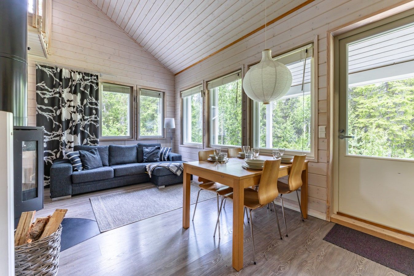 Une pièce chaleureuse avec un plafond en bois, une table à manger avec des chaises, un canapé et de grandes fenêtres donnant sur un paysage verdoyant.