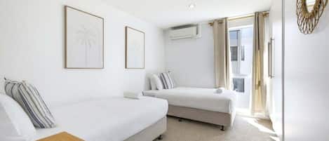 A hotel room with two single beds, a wooden side table, a white wall with two framed pictures, a window with curtains, and an air conditioning unit.