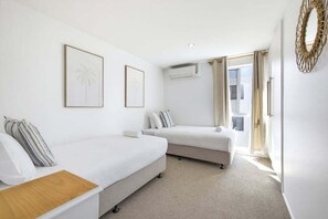 Ein Hotelzimmer mit zwei Einzelbetten, einem Holz-Nachttisch, einer weißen Wand mit zwei gerahmten Bildern, einem Fenster mit Vorhängen und einer Klimaanlage.