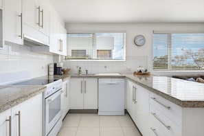 A modern kitchen with white cabinetry, a built-in oven, a dishwasher, and a window with blinds. - 11 Muroy Court - 50 Alexandra Parade Maroochydore (Maroochydore)