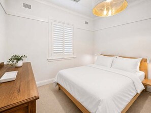 Ein Schlafzimmer mit Bett, Nachttisch, Lampe, Fenster mit Jalousien und einer Topfpflanze.