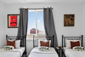 A room with two beds, a window with curtains, a framed red life jacket, and a painting on the wall.