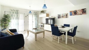 A modern living room with a sofa, a glass-top coffee table, and a dining area with a white table and blue chairs. - Casa Amadeus Corralejo (Corralejo)