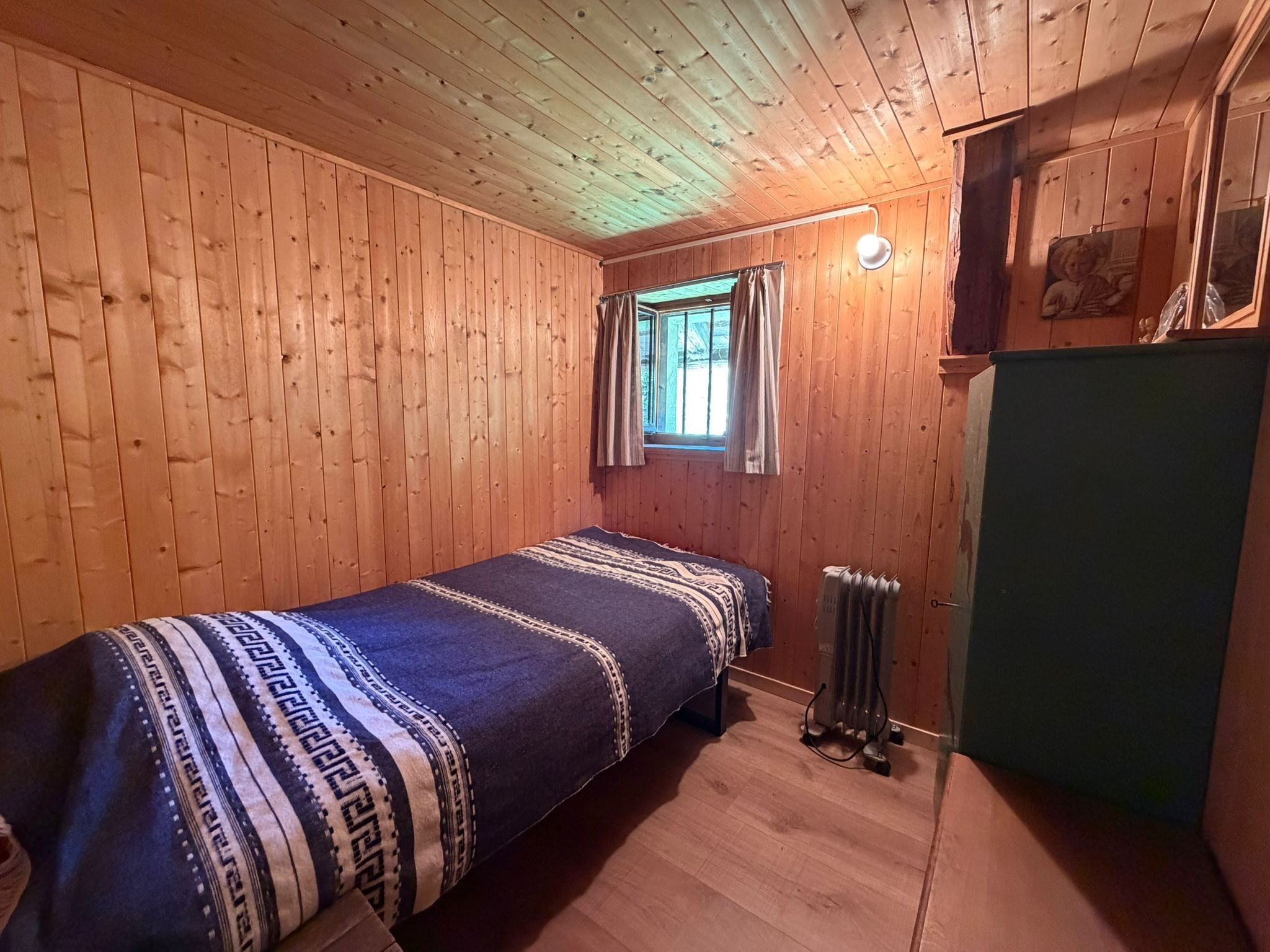 A small, single-bed room with wooden walls, a window with curtains, a radiator, and a green cabinet.