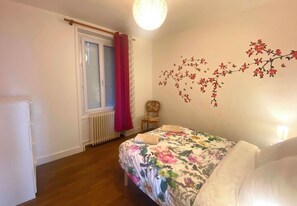 Un dormitorio con colcha estampada, un radiador, una silla y una ventana con cortinas.