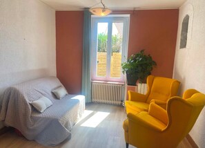 A room with a bed, a yellow armchair, a potted plant, and a window with a view of greenery.