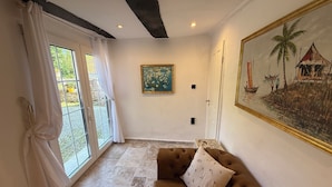 A room with a brown sofa, a painting of a coastal scene, a window with curtains, and a tiled floor.