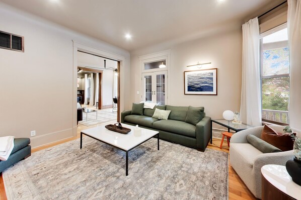 A living room with a green sofa, a white coffee table, and a grey sofa. There is a window with curtains, a framed picture on the wall, and a door leading to another room.