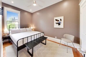 Un dormitorio con cama, una banca, una silla, una ventana y una obra de arte colgada en la pared.