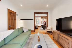 A living room with a green sofa, a wooden coffee table, a TV, and a view into the dining area. - The Reunion House: Two Massive Units in One! (Michigan City)