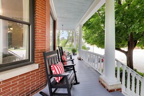 A covered porch with benches and a railing, a brick wall, and a tree. - The Reunion House: Two Massive Units in One! (Michigan City)