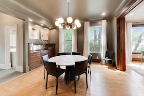 A dining area with a round table, chairs, a chandelier, and large windows with curtains. - The Reunion House: Two Massive Units in One! (Michigan City)