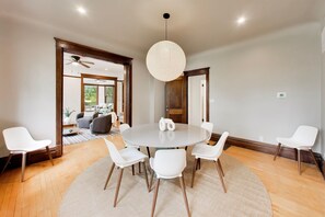 A dining area with a round table, white chairs, a hanging light fixture, and a view into a living room with a ceiling fan. - The Reunion House: Two Massive Units in One! (Michigan City)