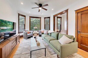 A spacious living room with a large flat-screen TV, a ceiling fan, and a large window providing a view of the outdoors.