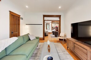 A living room with a green sofa, a wooden coffee table, a TV, and a view into the dining area. - The Reunion House: Tree Tops Flat Unit! (Michigan City)