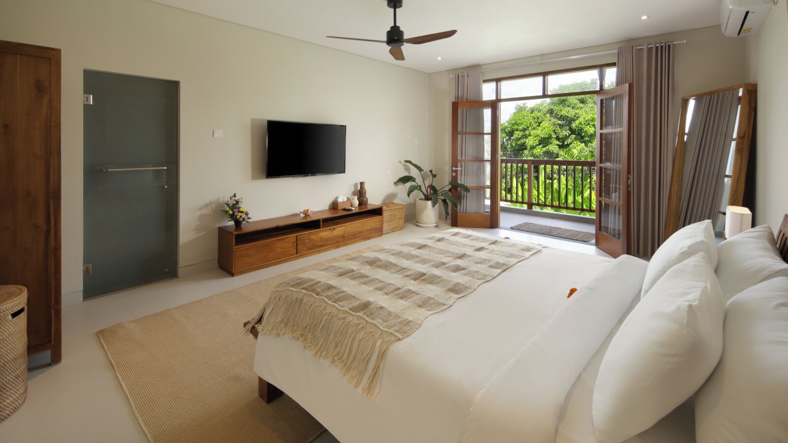 Quarto com cama, televisão, cómoda de madeira, varanda com cortinas e ventoinha no teto.