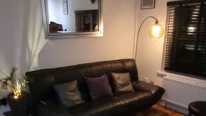 Uma sala de estar com um sofá de couro preto, uma mesinha branca, um abajur e um vaso com planta.