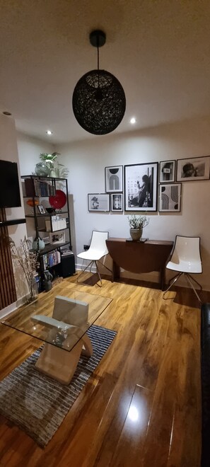 A modern living room with a glass coffee table, wooden flooring, a black ceiling light, a bookshelf, and framed pictures on the wall.
