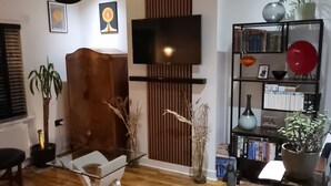 Sala de estar moderna com piso de madeira, mesa de centro de vidro, sofá de couro preto, TV fixada na parede e estante com diversos objetos.