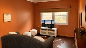 A room with an orange wall, a window with a view, a television, a white cabinet, and a dark-colored sofa. - Nandi - Vintage Character (Orange)
