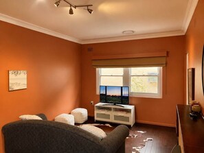 Ett rum med en orange vägg, ett fönster med utsikt, en tv, ett vitt skåp och en soffa i mörk färg.