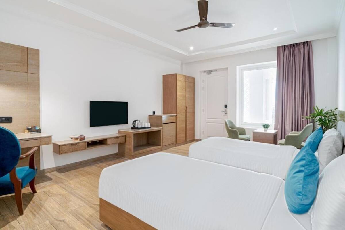 Habitación de hotel con cama, escritorio con televisor de pantalla plana, armario, silla, mesa pequeña y ventana con cortinas.