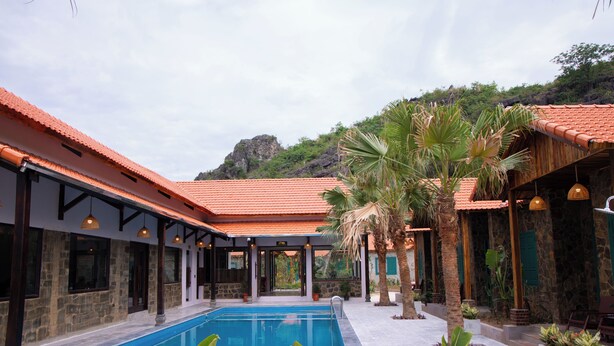 A swimming pool with a tiled deck, surrounded by stone walls and palm trees, with a mountain in the background. - Laco green Trang An (Tay Hoa Lu)