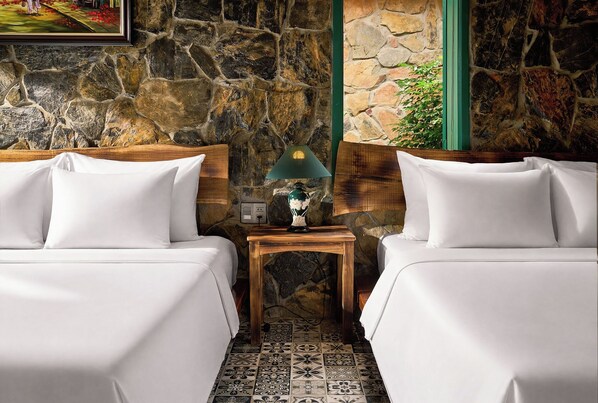 A hotel room with two beds, a nightstand with a lamp, and a stone wall.