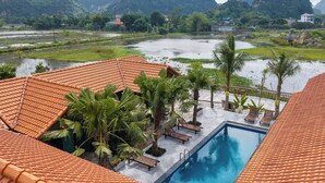 A swimming pool with a tiled roof, surrounded by palm trees and lounge chairs, overlooking a flooded landscape with buildings in the distance.