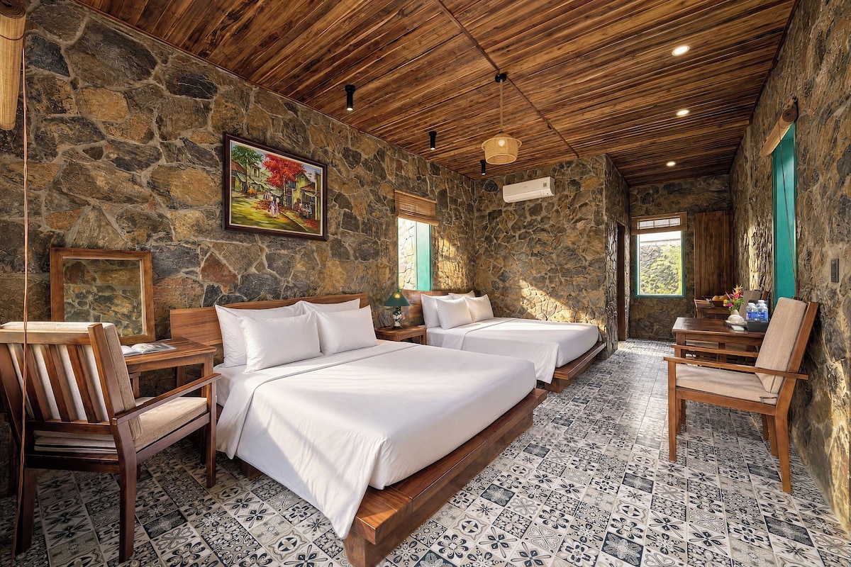 A room with a stone wall, wooden ceiling, two beds, a chair, a table, and a painting.