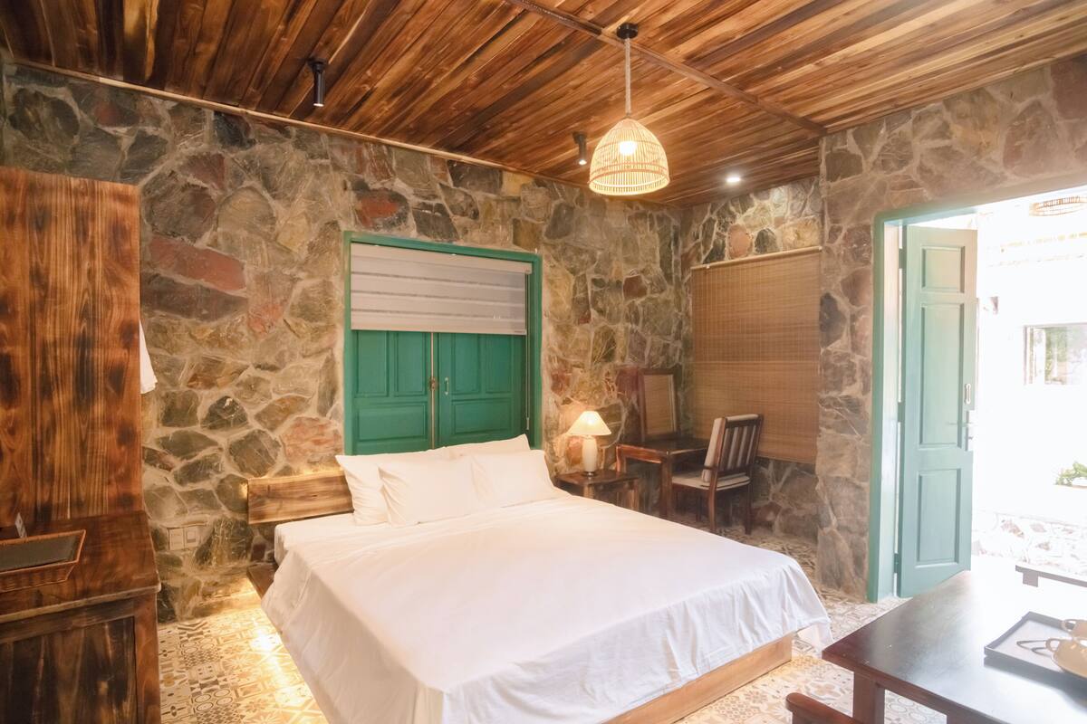 Una habitación con una cama, un escritorio de madera, una silla, una lámpara y una pared de piedra.