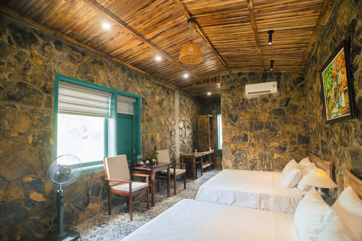 Una habitación con paredes de piedra, techo de madera, dos camas, una mesa con sillas, un ventilador y una ventana con persianas.