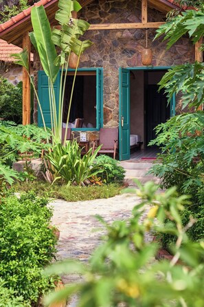 A stone building with two open doors, a wooden porch, and a garden path leading to a seating area.