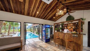 A bar area with a wooden counter, stools, and a Pepsi refrigerator. There is a pool visible through an open door.