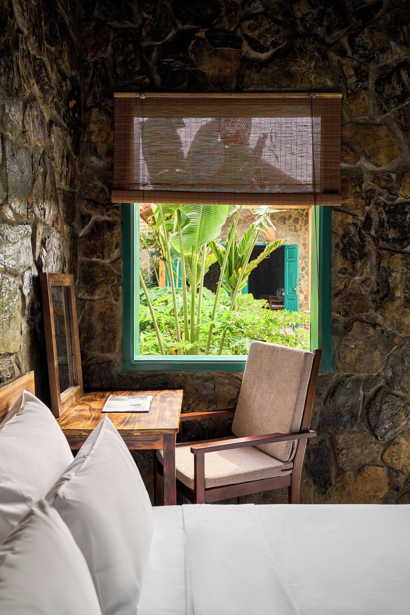 A room with a stone wall, a window with a view of greenery, a wooden table, a chair, and a bed with white bedding.