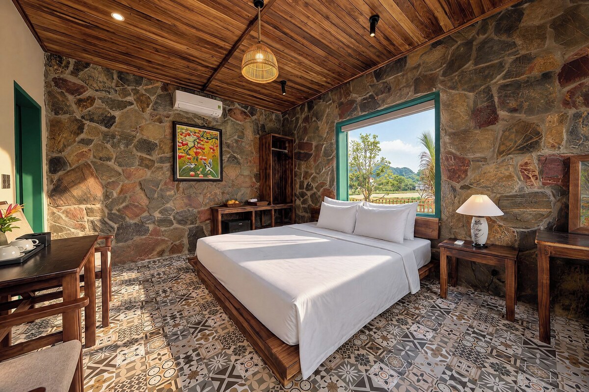 A bedroom with a stone wall, a bed with white linens, a wooden desk, a lamp, and a view of the outdoors.