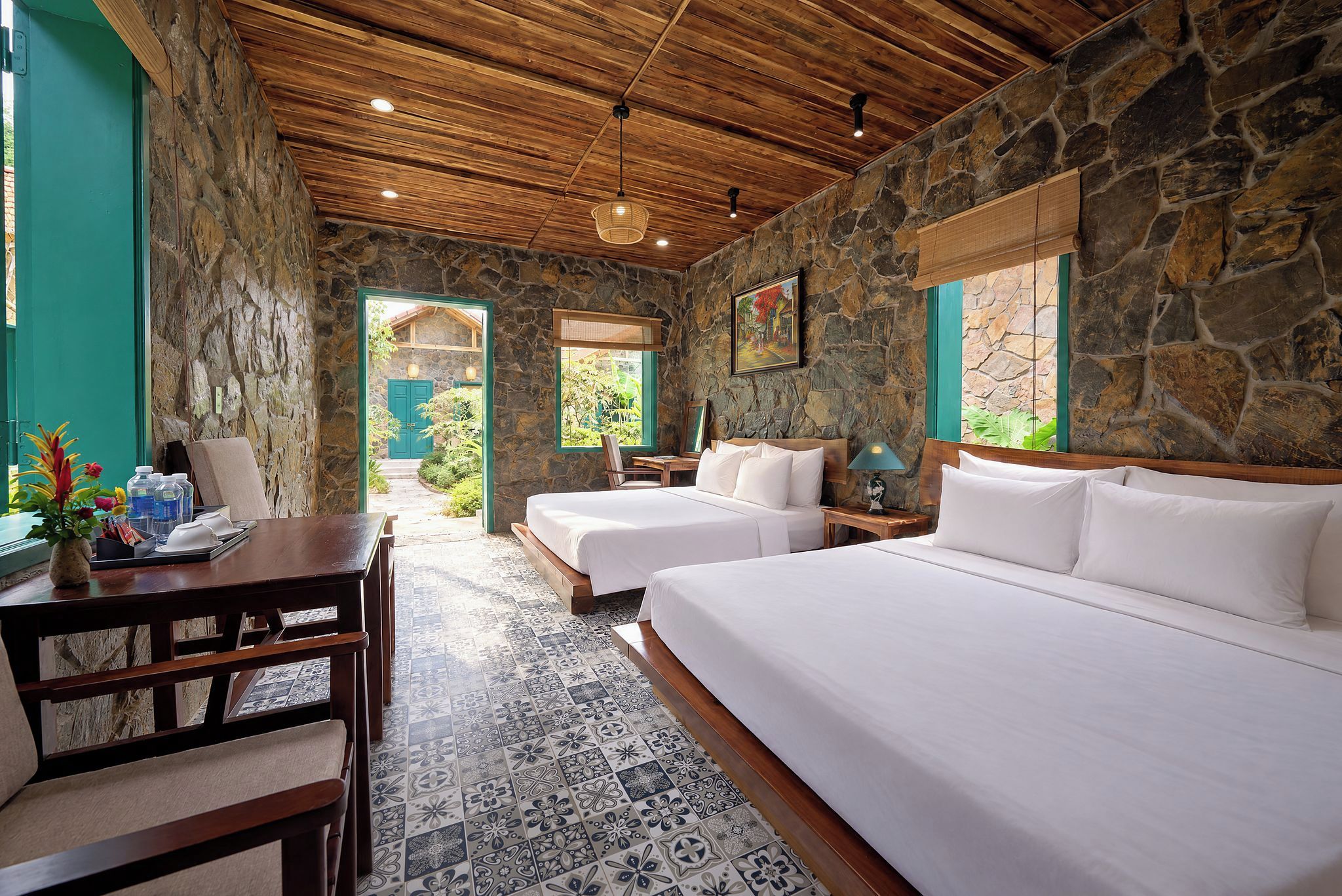 A room with a stone wall, wooden ceiling, two beds, a desk, and a view of a garden.