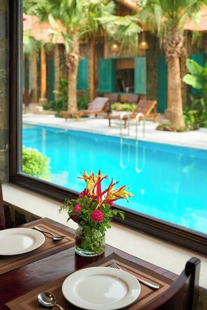 A dining table with a vase of flowers and plates, overlooking a swimming pool with lounge chairs and palm trees.