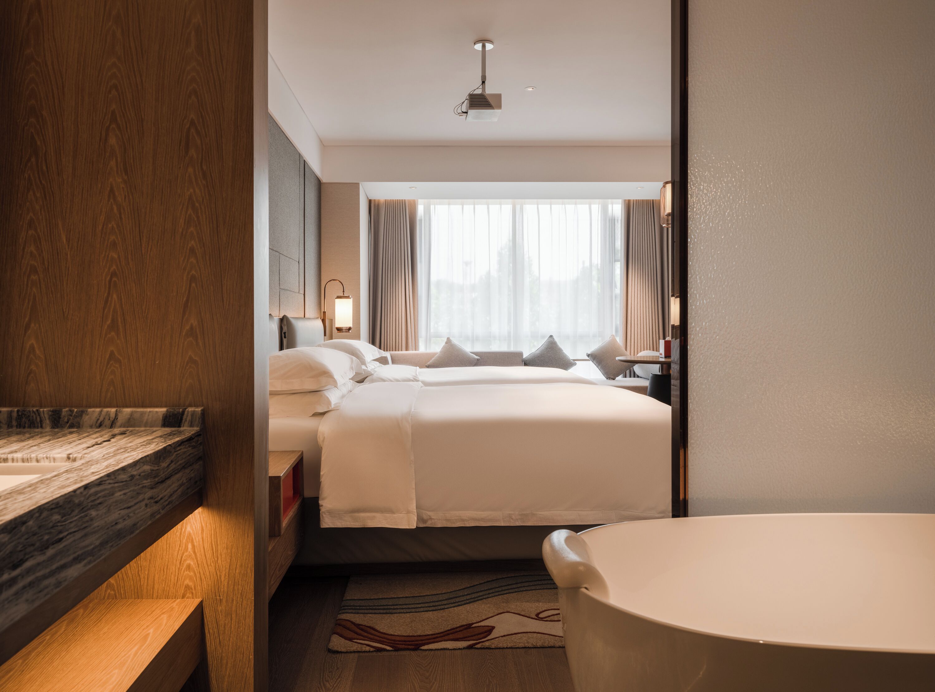 Habitación de hotel con cama, mesita de noche, lámpara y vistas a la ciudad desde la ventana.