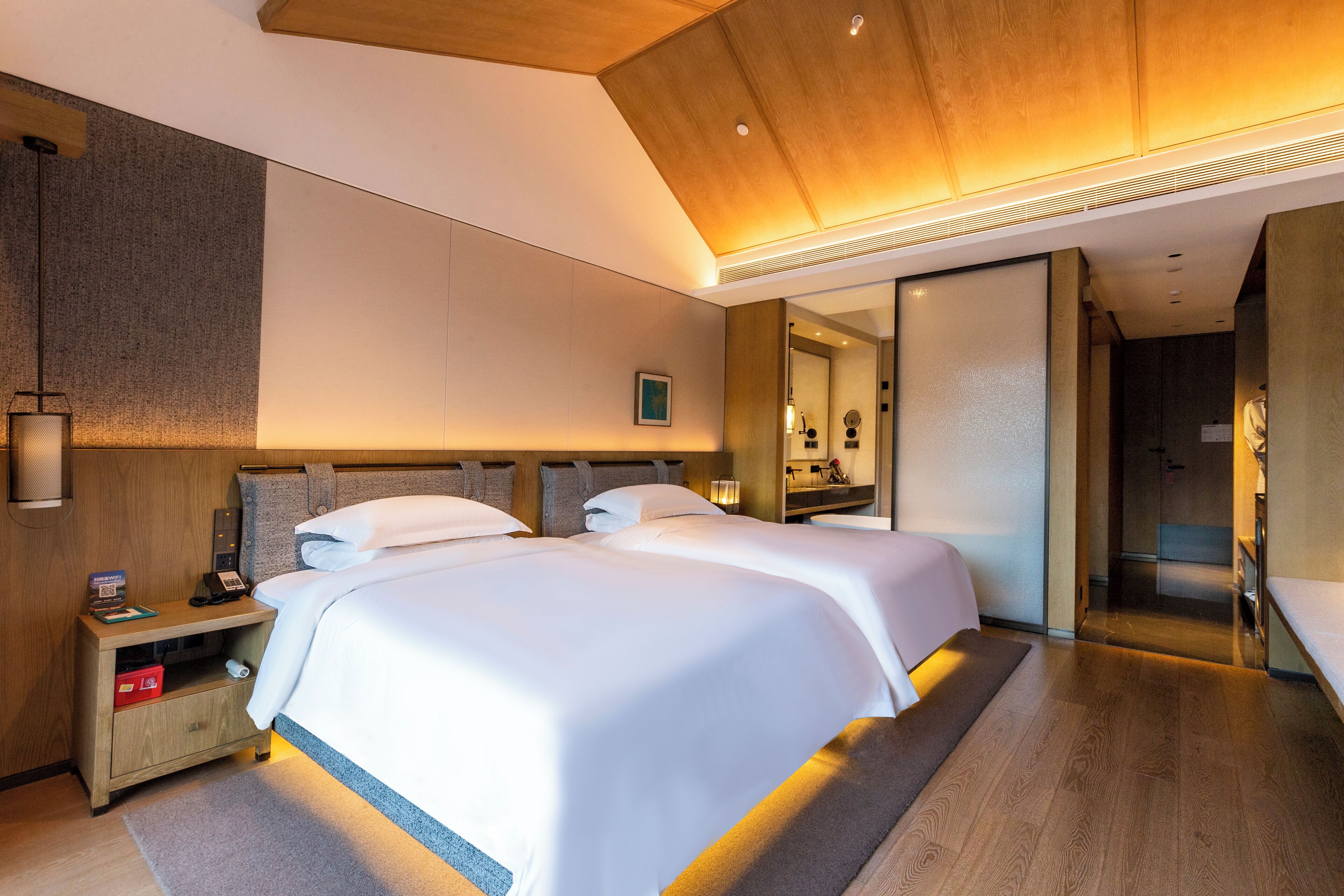 Een moderne hotelkamer met twee bedden, een nachtkastje, een spiegel en een houten plafond.